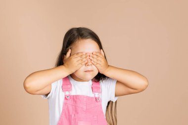  Çocuk elleri kapalı gözlerini kapatıyor. Diğerlerini ağzı kapalı görmüyor. Diğerleriyle parlak pembe tulum ve bej arka planda beyaz tişört giyerek konuşmuyorlar.