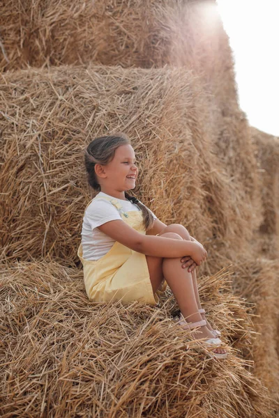 Portrait of sitting girl on high haystack smiling with teeth looking somewhere far away wearing sundress. Having fun away from city on field full of golden hay — Stock Photo, Image