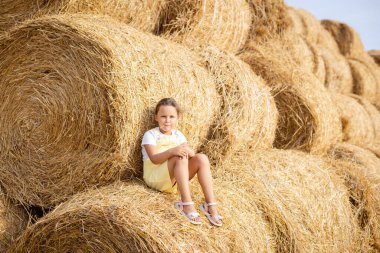 Portrait of shining happy small girl with piece of hay in mouth in sundress resting on tall haystack and leaning on another stack and other haystacks in background. Field full of golden hay
