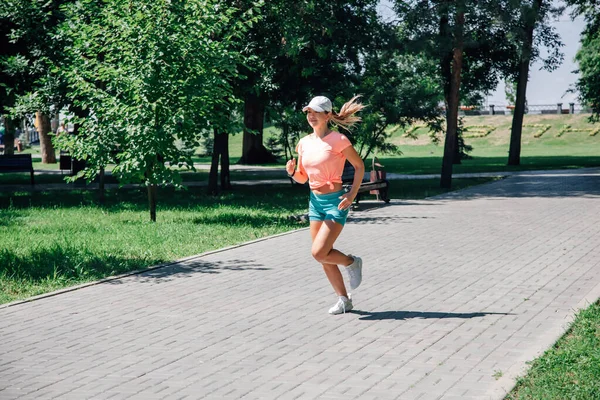 Running smiling young sporty woman in park in sportswear in front of green trees on road of grey tiles in sunny summer day — Stock Photo, Image