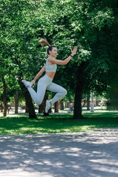 Smiling young sporty woman jumping in air stretching out arms joyfully in grey sportswear in summer sunny day in park — Stok fotoğraf