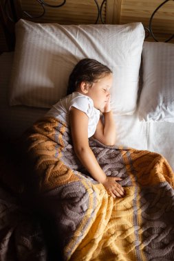 Young female kid sleeping and having rest on left side with one hand under head on big double bed covered with yellow gray blanket wearing home clothes