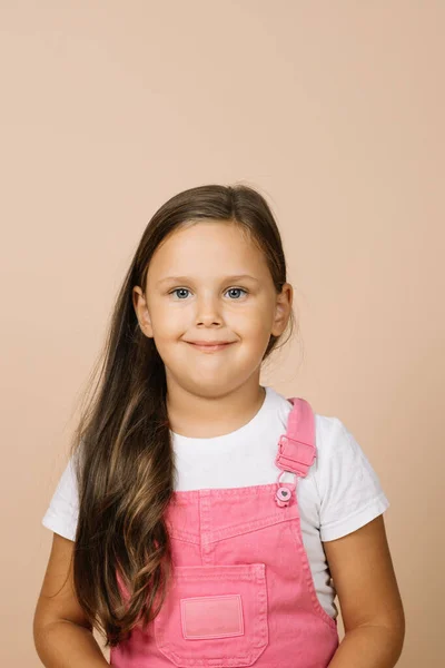 Close up face kid with bright shining eyes and calm happy wide smile looking at camera wearing bright pink jumpsuit and white t-shirt on beige background — Fotografia de Stock
