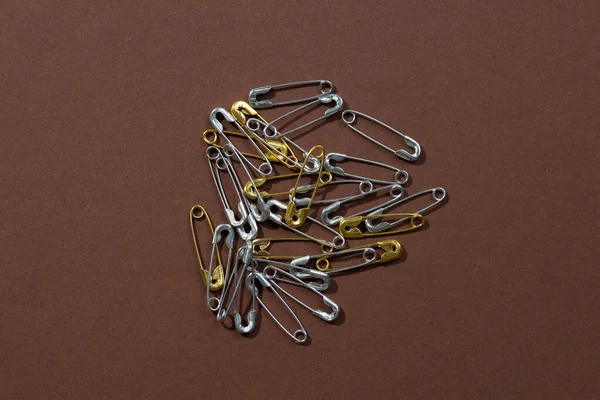 Dark-brown background with tiny silver and gold safety pins scattered chaotically in center. Copy space. Mock up photography. Creative minimalism with office stationery — Foto de Stock