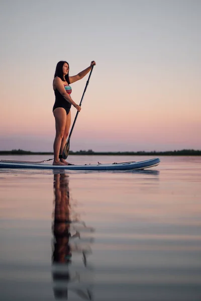 Porträtt av medelålders kvinna pöl ombordstigning med pöl i händerna tittar på solnedgången på sjön med spegel reflektion i vatten på kvällen med rosa himmel i bakgrunden. Aktiv livsstil för äldre — Stockfoto