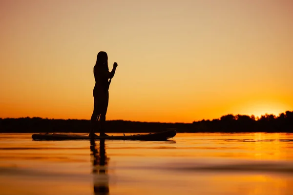 傍晚时分，女人手握桨，从低矮的角度向水面上射击，凝视着覆盖着橙色水面的令人惊奇的落日。积极的生活方式 — 图库照片