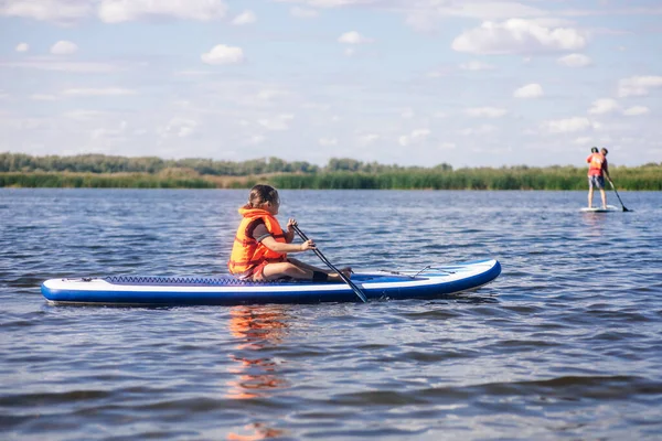 一个白人小女孩一个人坐在桨板上，手里拿着桨，看着其他穿着橙色救生衣的潜水员。积极的假期。从小就灌输对体育运动的热爱 — 图库照片