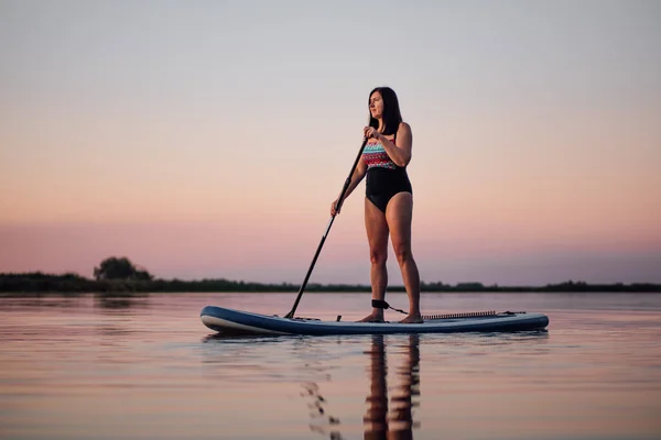 Entspannt rudert eine Frau mittleren Alters auf einem Surfbrett mit Ruder und blickt auf den Sonnenuntergang auf dem See, dessen Wasser mit rosa Himmel im Hintergrund plätschert. Aktiver Lebensstil für ältere Menschen — Stockfoto