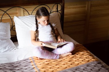 Küçük ciddi kız kitap okuyor, dikkatle yatakta oturuyor ve ahşap evde ev kıyafetleri giyiyor. Boş zaman aktiviteleri. Kendi kendini geliştirme