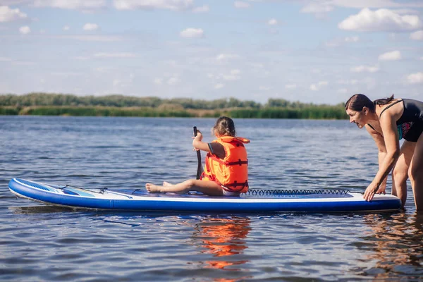 Mãe ajudando a filhinha a comer a bordo com remo nas mãos no lago com juncos verdes e árvores no fundo vestindo colete colete salva-vidas. Férias activas. Inculcação de amor por esportes desde a infância — Fotografia de Stock