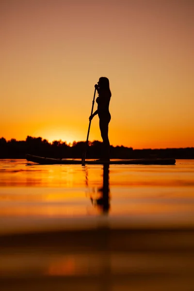 女子划桨在海湾上的低角度俯瞰着水面，在夕阳的映照下思考着身后的事。室外积极的生活方式 — 图库照片