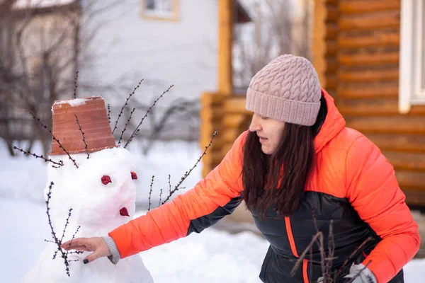 Μεσήλικη γυναίκα μόνη της κάνει χιονάνθρωπο στην πίσω αυλή του εξοχικό σπίτι κατά τη διάρκεια της ημέρας, ενώ το χιόνι πέφτει κάτω με ξύλινο σπίτι στο παρασκήνιο. Γονείς που περνούν χρόνο με παιδιά — Φωτογραφία Αρχείου