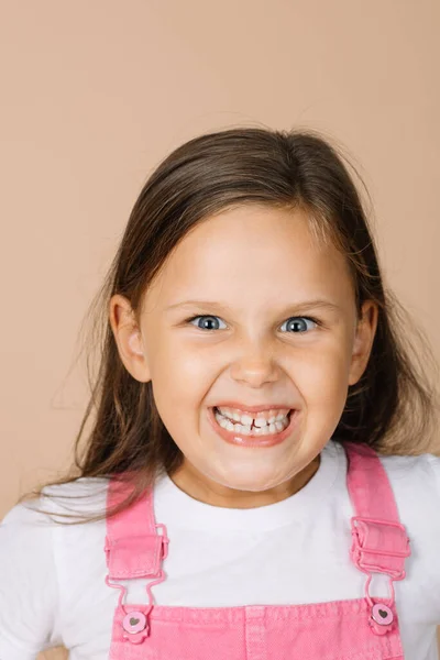 Retrato de niña con aterradora sonrisa aterradora sonrisa forzada con todos los dientes con ojos brillantes juguetones mirando a la cámara con mono de color rosa brillante y camiseta blanca sobre fondo beige — Foto de Stock