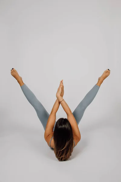 Brunette está envolvida em esportes. Mulher jovem realiza exercício de ioga ou fitness, ela se deita de costas, abriu as pernas levantadas para os lados, cruzou os braços em suas palmas das mãos, visão traseira, fundo isolado — Fotografia de Stock