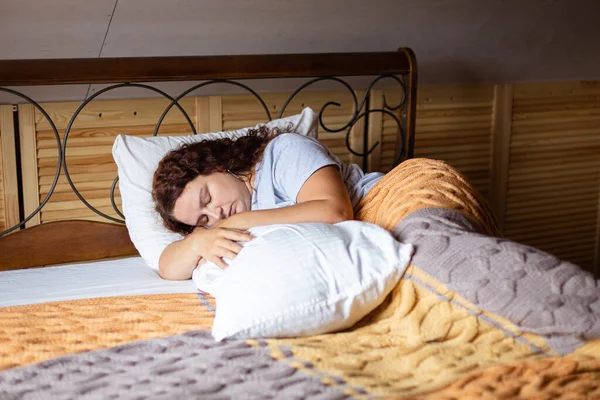 Расстроенная кудрявая брюнетка-взрослая женщина в постели под махровыми одеялами с закрытыми глазами, обнимающая подушку в домашних пижамах днем. Тревожный момент во взрослой жизни — стоковое фото