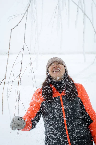 Lächelnde Frau mittleren Alters, die mit Schnee bedeckte Zweige zieht, warm angezogen auf schneebedecktem Boden steht und aufblickt, während Schnee auf sie fällt. Schöne Winterzeit voller weißer Farben — Stockfoto