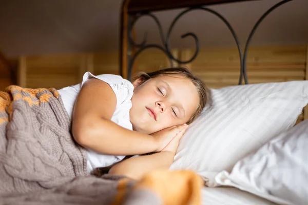 Detailní záběr holčičky spící na velké posteli s oběma rukama pod hlavou přikrytýma dekou v domácím oblečení v dřevěném domě. Spát ráno o víkendu — Stock fotografie