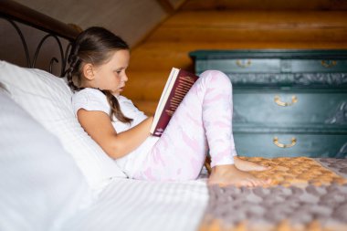 Kafkasyalı hevesli bir çocuk, elinde kitapla büyük bir dikkatle yatağa uzanmış, ahşap evde pijamalarıyla kitap okuyor. Boş zaman aktiviteleri. Kendi kendini geliştirme