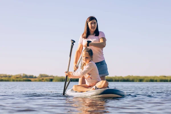 中年妇女和女儿划船时，努力地坐在冲浪板上，与绿芦苇和绿树一起锻炼和享受时光。积极的生活方式。家人在一起的时间 — 图库照片