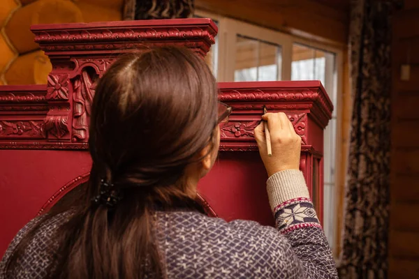 Sorgfältiger Prozess der Möbelpatinierung. Frau mit Pinsel, roter Holzschrank mit brillanten geschnitzten Ornamenten. Umweltfreundliche Wiederverwendung. Alte Dinge bekommen neues Leben lizenzfreie Stockbilder