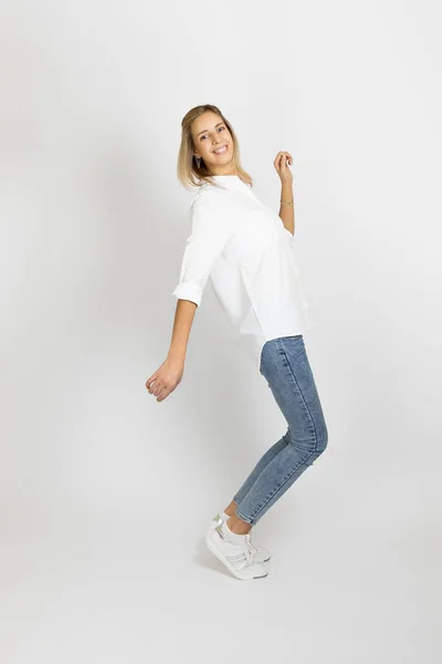Сияющая молодая женщина с улыбкой на лице позирует на белом фоне студии, глядя на камеру в белой блузке, синих джинсах и кроссовках. Показать положительные эмоции — стоковое фото