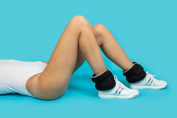 Fit jonge vrouw met lange benen dragen bodysuit en sneakers met enkel gewichten op blauwe achtergrond doen fysieke oefeningen. Gezonde levensstijl, trainen — Stockfoto