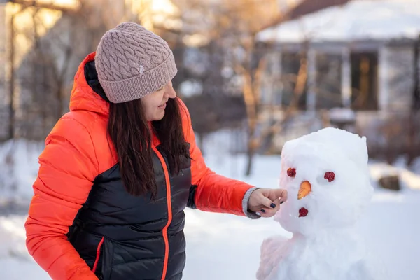 Vit medelålders kvinna gör snögubbe ansikte med morot och kottar på dagtid samtidigt som du har promenader i parken med träd, hus och starkt solljus i bakgrunden. Föräldrar som umgås med barn — Stockfoto