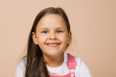 Parlak gözlü çocuk portresi ve parlak pembe tulum ve bej arka planda beyaz tişört giyen kameraya bakan üst dişli mutlu gülümseme.