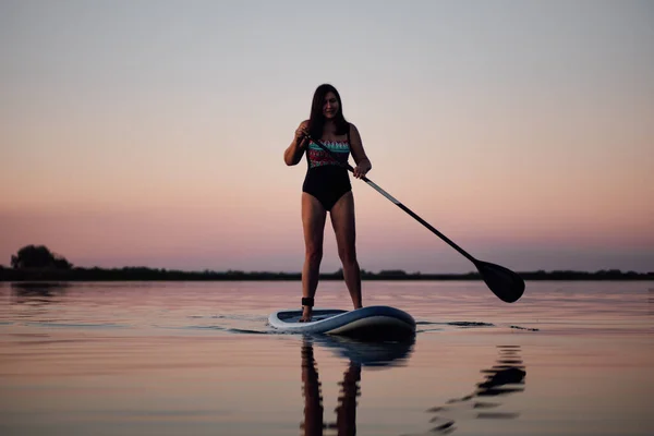 夏天，雌性潜水员手握桨划船，看着镜头在平静的湖面上，背景是迷人的粉色天空，身穿泳衣。老年人的积极生活方式 — 图库照片