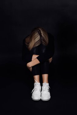 Desperate blonde woman with face put on knees sitting on floor and hiding from people on black background. Physical and mental abuse. Relative aggression clipart