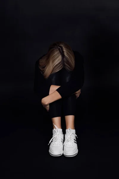 Desperate blonde woman with face put on knees sitting on floor and hiding from people on black background. Physical and mental abuse. Relative aggression