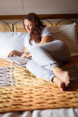 Kıvırcık esmer kadın yetişkin terry yatak örtüsüne uzanmış, elinde kitap tutan mutlu yüzüyle gündüzleri evde pijama giyiyordu. Rahatlatıcı boş zaman aktivitesi