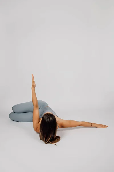 A mulher pratica desporto. Jovem morena realiza exercício de ioga ou fitness, ela se deita de costas e levanta um braço, pernas dobradas e colocadas no chão, fundo isolado cinza, visão traseira — Fotografia de Stock