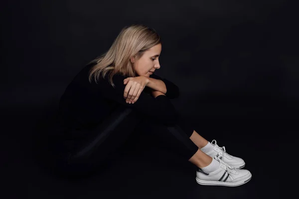 Caucasian Blonde self-contained woman closing herself from other people sitting on floor with black background. Domestic physical and psychological abuse — Stock Photo, Image