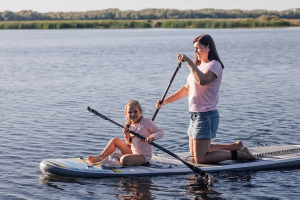 Medelålders mamma lär liten kvinnlig unge att göra pöl ombordstigning på sjön med gröna träd och vass i bakgrunden. Aktiv livsstil. Att lära barn att älska idrott från tidig ålder — Stockfoto