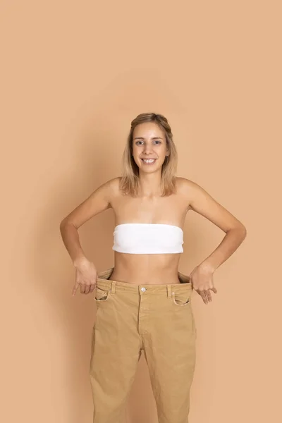 Retrato de mujer sonriente en buena forma en pantalones de gran tamaño, ropa interior blanca que muestra la cintura delgada tirando de los pantalones en el fondo de color de la piel. Perder peso. Mantener una dieta saludable, hacer ejercicio — Foto de Stock