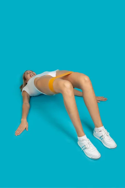 Sportieve vrouw die traint met een verzetsband in wit bodysuit en sneakers op een turquoise achtergrond. Sporten om in goede conditie te zijn. Gezondheid thuis verbeteren — Stockfoto