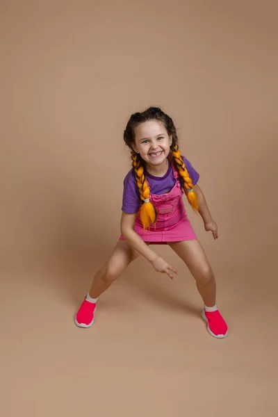 Niña activa feliz que tiene trenzas de kanekalon amarillo bailando con las piernas separadas sonriendo mostrando el signo de la pieza con la mano en mono rosa, camiseta púrpura y zapatillas de deporte de color rosa sobre fondo beige — Foto de Stock
