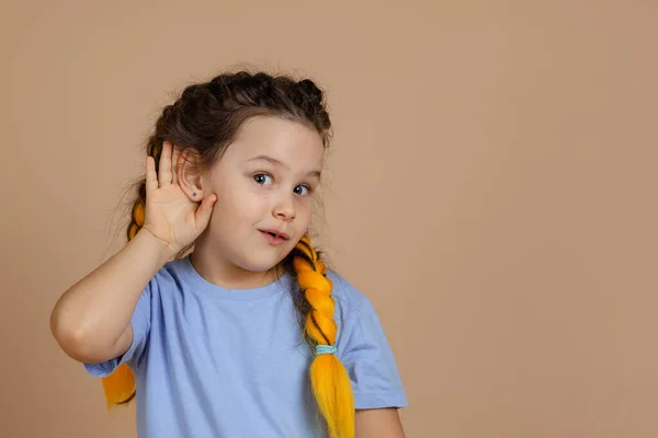 Neugierig interessiertes kleines kaukasisches Mädchen mit Kanekalon-Zöpfen mit leuchtenden Augen, die Ohren halten, um etwas zu überhören, das auf beigem Hintergrund in die Kamera schaut Stockbild