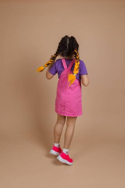 İki kanekalon sarı örgülü, pembe tulum, mor tişört ve bej arka planda pembe spor ayakkabılar giyen enerjik, küçük bayan bir çocuk.