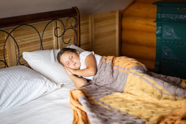 Młoda biała dziewczyna drzemie na wielkim podwójnym łóżku z obiema rękami pod głową pokrytymi kocem z komodą za łóżkiem w drewnianym domu. Spanie rano w weekend — Zdjęcie stockowe
