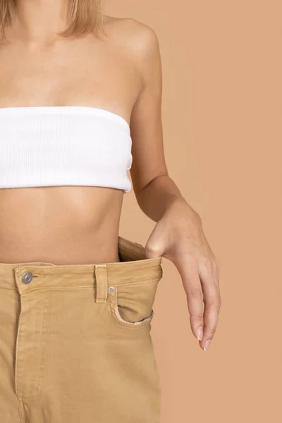 Κοντινό πλάνο της λεπτής Καυκάσιας γυναίκας που φοράει υπερμεγέθη παντελόνια και λευκά εσώρουχα σουτιέν που δείχνουν λεπτή μέση στο χρώμα του δέρματος φόντο. Απώλεια βάρους. Διατήρηση υγιεινής διατροφής, άσκηση — Φωτογραφία Αρχείου