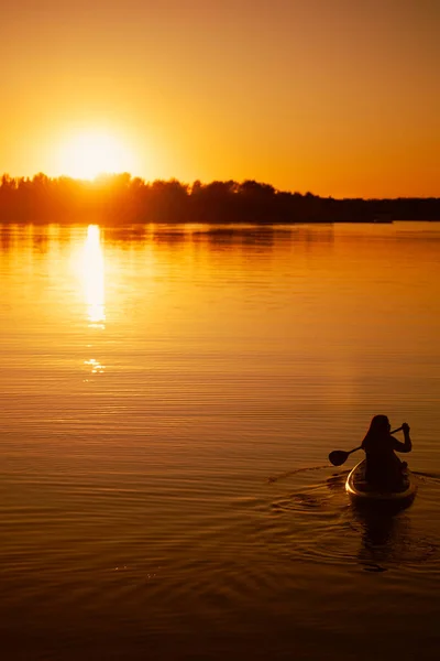 Silhouette of woman sitting on paddle board with oar with hands rowing straight on beautiful lake with astonishing sunset in background mirroring in water — Stock Photo, Image