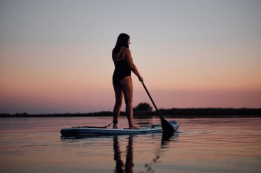 Arka planda muhteşem pembe gökyüzü ve mayo giymiş suya bakan kürekli orta yaşlı bir kadının arkası. Yaşlılar için aktif yaşam tarzı