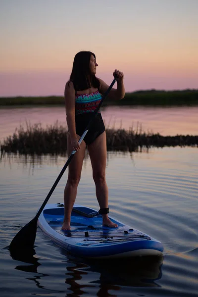 中年妇女手握桨，望着蓝色的冲浪板，在晚霞的湖畔，背景是美丽的粉红天空。老年人的积极生活方式 — 图库照片