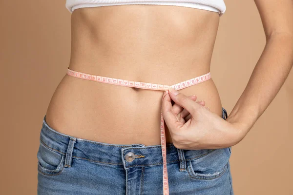 Fêmea com fita mostrando resultado de perda de peso medir a cintura em sutiã branco e jeans azul no fundo bege. Manter a dieta e o exercício. Estabelecimento e realização de metas — Fotografia de Stock