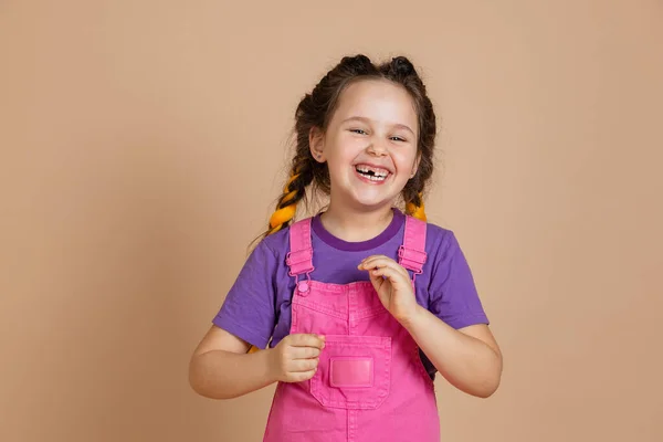 Porträt einer lachenden kleinen Frau mit Kanekalon-Zöpfen und fehlenden Zähnen, die in die Kamera blickt und einen rosafarbenen Overall und ein lila T-Shirt auf beigem Hintergrund trägt Stockfoto
