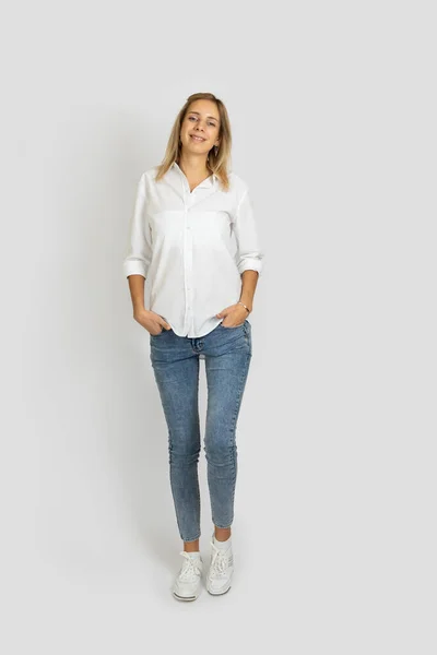 Mujer alta en forma de pie sobre fondo blanco estudio mirando a la cámara posando con las manos en bolsillos en blusa blanca, vaqueros azules y zapatillas de deporte. Mostrando emociones positivas — Foto de Stock