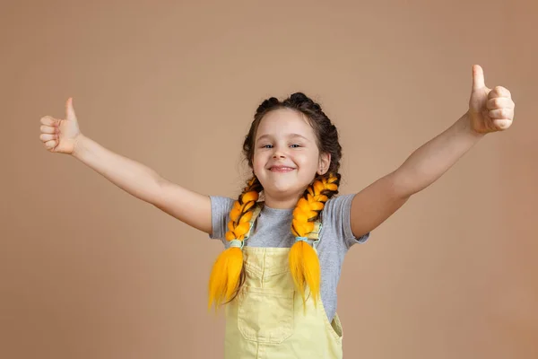Joven niña sonriente feliz con coletas de kanekalon amarillo mirando a la cámara, mostrando como signos con las manos usando mono amarillo y camiseta gris sobre fondo beige — Foto de Stock