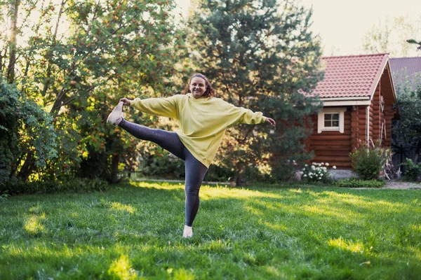 Kobieta dorosła uśmiechnięta na zielonej trawie ćwiczy w odzieży sportowej z jedną nogą na podwórku pełnym światła słonecznego z domkiem na wsi i wysokimi drzewami w tle. Pozytywny wynik — Zdjęcie stockowe